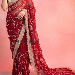 May include: A deep red saree featuring intricate gold floral embroidery and a wide, decorative gold border with fringe detailing. The garment is paired with a sleeveless, cream-colored blouse adorned with gold embroidery and a scoop neckline.