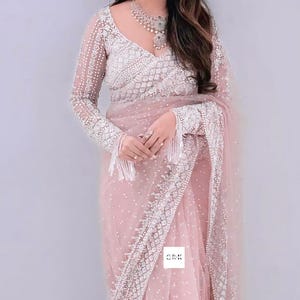 Può includere: Un sari rosa chiaro con ricami e abbellimenti bianchi intricati. Il sari presenta un tessuto trasparente e puntinato e maniche lunghe con frange. Una collana abbinata completa l'elegante design.
