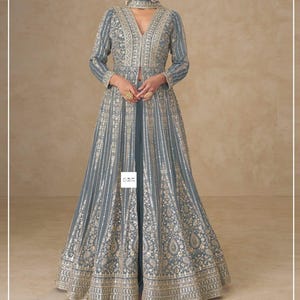 Può includere: Un lungo abito grigio fluente con intricati ricami dorati. L'abito presenta uno scollo a V, maniche lunghe e un choker abbinato. Il design include strisce verticali e motivi floreali, creando un look lussuoso ed elegante. Adatto per eventi formali.