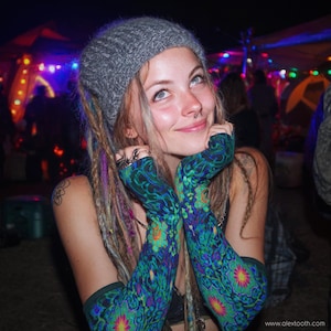 May include: A person wearing a gray beanie, patterned arm warmers, and dreadlocks. The arm warmers feature a blue, green, and orange floral design. The background is blurred with colorful lights.