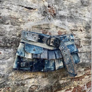 Puede incluir: Minifalda vaquera con diseño desgastado y en capas. La falda presenta un cinturón ancho con una hebilla grande y ojales plateados. La falda está colgada en una percha de plástico transparente contra una pared de ladrillo desgastada.