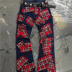 Puede incluir: Vaqueros de mezclilla azul oscuro con parches de cuadros rojos y negros y bordes deshilachados. Los vaqueros tienen un cinturón de cuero negro con tachuelas piramidales plateadas y imperdibles plateados. Un artículo de moda único, de inspiración punk.