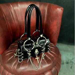 Könnte beinhalten: Schwarze Handtasche aus Lackleder mit silberfarbener Hardware. Die Tasche hat zwei obere Griffe, große Metallringe und dekorative Kreuzanhänger. Sie hat eine rechteckige Form mit einem Klappenverschluss.