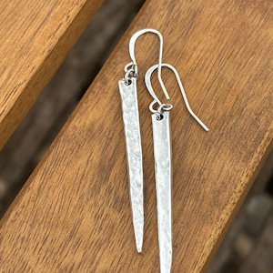 May include: A pair of silver-colored, hammered metal earrings. Each earring has a long, rectangular shape that tapers to a point. The earrings hang from a simple hook. The background is a wooden surface.