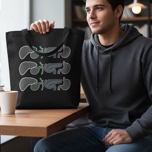 May include: Black tote bag featuring a repeating pattern of stylized internal organs in gray and light green. The bag is held by a person. A white cup and books are on a wooden table.