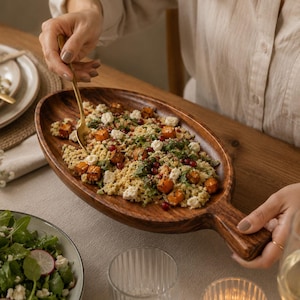 May include: An oval-shaped wooden serving platter with a handle, featuring a natural dark brown wood grain. The platter is filled with a salad containing couscous, roasted orange squash cubes, white cheese crumbles, and red pomegranate seeds.