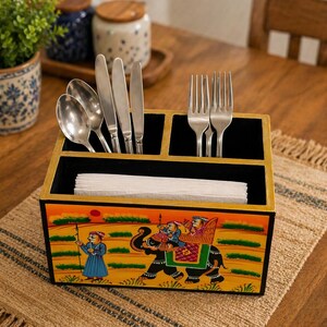 Puede incluir: Un portacubiertos de madera pintado a mano con un diseño de elefante y figuras con vestimenta tradicional. El soporte tiene compartimentos para cubiertos y servilletas. El soporte está sobre una mesa de madera.