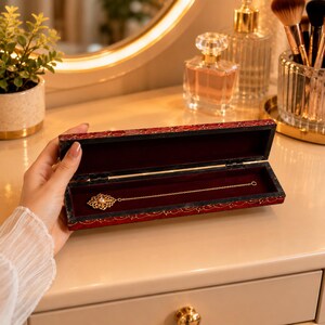 Pode incluir: Um colar dourado com um pendente decorativo é exibido dentro de uma caixa de joias com padrão vermelho e preto. A caixa está aberta, revelando um interior de veludo. O colar está sobre uma superfície branca.