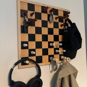 May include: A wall-mounted organizer featuring a black and light wood checkerboard pattern. The board includes several protruding wooden chess pieces in dark brown and white, serving as hooks for hanging items like keys, a black cap, and headphones.