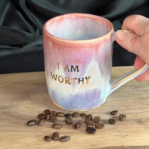 May include: A ceramic mug with a pink, purple, and white glaze, featuring the gold text "I AM WORTHY". Coffee beans are scattered on a wooden surface near the mug. The mug has a handle and is held by a person.