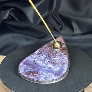 Könnte beinhalten: Ein tropfenförmiger Räucherstäbchenhalter mit lila und brauner Glasur. Der Keramikhalter hat ein geprägtes Blumendesign und eine goldfarbene Metallkugel, um den Räucherstäbchen zu halten. Der Räucherstäbchen ist angezündet und gibt Rauch ab.