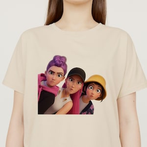 May include: Beige t-shirt featuring a graphic of three animated female characters with expressive faces. One has purple hair, another wears a black cap, and the third has a yellow hat. The characters are rendered in a cartoon style.