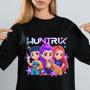 May include: Black long-sleeve shirt featuring the word "HUNTRIX" in a blue and purple gradient. The shirt has a graphic of three cartoon characters with different hair colors and outfits, surrounded by purple star shapes.