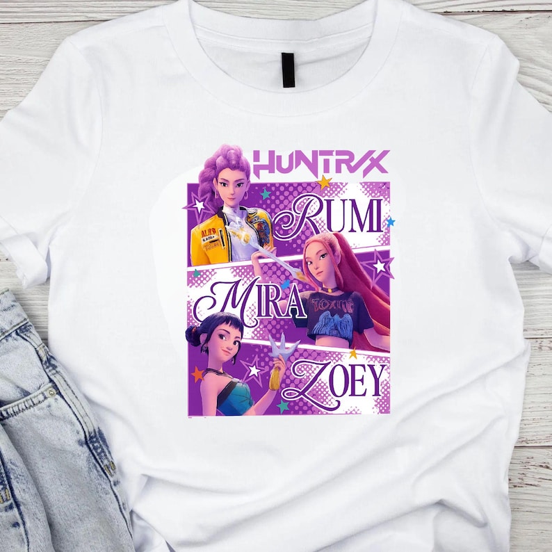 May include: White t-shirt featuring a graphic design with the word "HUNTRAX" above three illustrated female characters named Rumi, Mira, and Zoey. The design includes purple and yellow accents and star shapes.