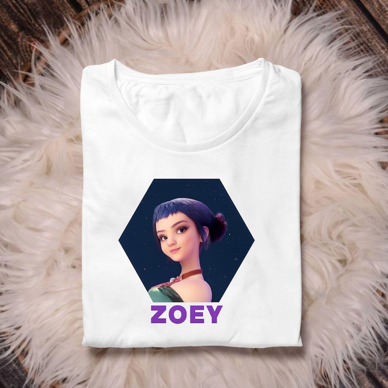 May include: White t-shirt featuring a graphic of a woman with blue hair and the name "Zoey" in purple text. The graphic is set within a dark blue hexagon. The shirt is displayed on a fluffy, white surface.