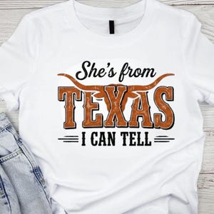May include: White t-shirt featuring the phrase "She's from TEXAS I CAN TELL" in a distressed brown and black design. The word "TEXAS" is in large, bold letters with a longhorn graphic above it.