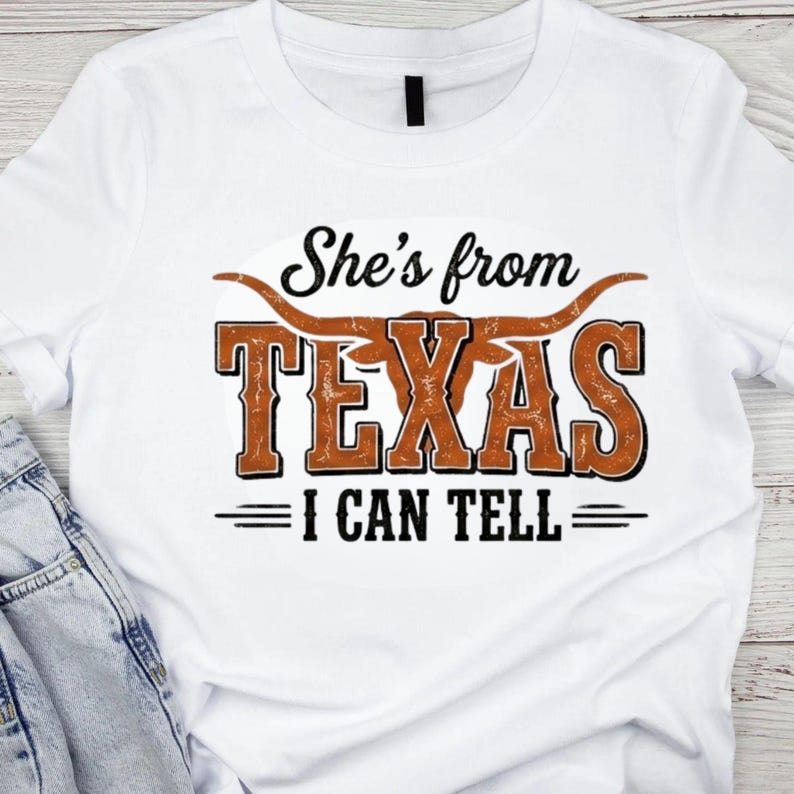 May include: White t-shirt featuring the phrase "She's from TEXAS I CAN TELL" in a distressed brown and black design. The word "TEXAS" is in large, bold letters with a longhorn graphic above it.