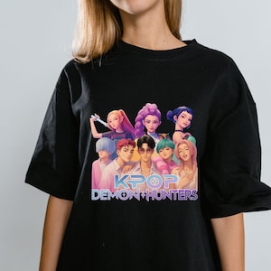 May include: Black t-shirt featuring a colorful graphic of a group of anime-style characters. The text "KPOP DEMON HUNTERS" is displayed in white and gold. The shirt is made of a soft material.