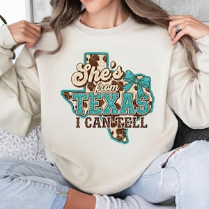 May include: Cream-coloured sweatshirt featuring a Texas-shaped graphic. The design includes the text "She's from TEXAS I CAN TELL" in brown, turquoise, and cowhide print. A turquoise bow is also featured.