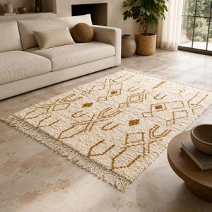 May include: A rectangular area rug featuring a cream-colored base with a repeating geometric pattern in mustard yellow. The rug has a textured, shaggy pile and is finished with knotted fringe detailing along the two shorter edges of the piece.