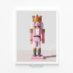 May include: A framed print of a pink nutcracker soldier with a gold crown, mustache, and black boots. The nutcracker is standing on a pink base. The print has the text "Pixel Print Shop Studio" at the bottom.