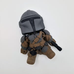 Puede incluir: Una peque&ntilde;a figura estilizada de un personaje con casco y armadura grises, sosteniendo un arma negra. La figura tiene detalles marrones y grises, con una placa de pecho y botas detalladas. La figura est&aacute; sobre una superficie blanca.