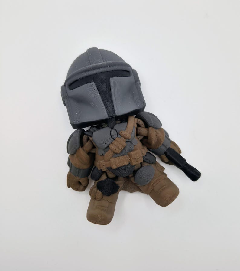Puede incluir: Una peque&ntilde;a figura estilizada de un personaje con casco y armadura grises, sosteniendo un arma negra. La figura tiene detalles marrones y grises, con una placa de pecho y botas detalladas. La figura est&aacute; sobre una superficie blanca.