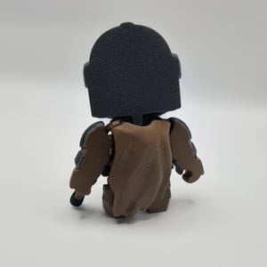 Puede incluir: Figura impresa en 3D de un personaje con casco negro y armadura marr&oacute;n. La figura est&aacute; en posici&oacute;n sentada, con una capa marr&oacute;n sobre la espalda. La figura tiene un aspecto texturizado.