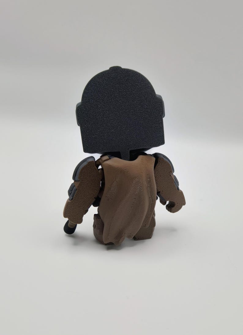 Puede incluir: Figura impresa en 3D de un personaje con casco negro y armadura marr&oacute;n. La figura est&aacute; en posici&oacute;n sentada, con una capa marr&oacute;n sobre la espalda. La figura tiene un aspecto texturizado.