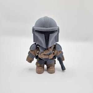 Puede incluir: Peque&ntilde;a figura de acci&oacute;n gris del Mandaloriano, con casco, armadura y bl&aacute;ster. La figura tiene detalles en marr&oacute;n y gris, y est&aacute; sobre una superficie blanca. Es un art&iacute;culo de colecci&oacute;n.