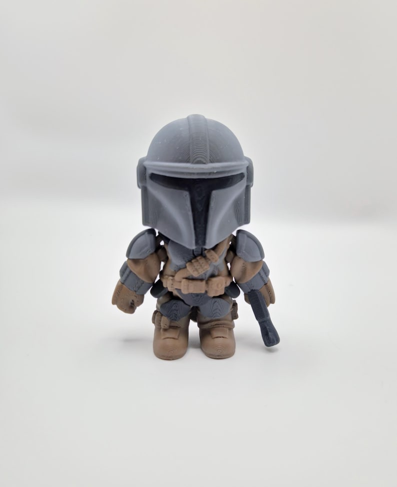 Puede incluir: Peque&ntilde;a figura de acci&oacute;n gris del Mandaloriano, con casco, armadura y bl&aacute;ster. La figura tiene detalles en marr&oacute;n y gris, y est&aacute; sobre una superficie blanca. Es un art&iacute;culo de colecci&oacute;n.
