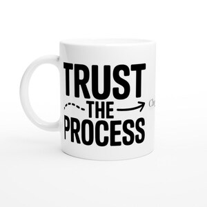 Op de afbeelding: Witte keramische mok met de woorden "TRUST THE PROCESS" in vette zwarte letters. Een stippellijn met een pijl wijst van "TRUST" naar "THE PROCESS". De mok heeft een gebogen handvat.