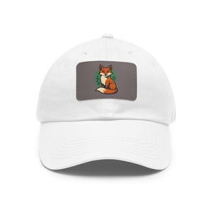 May include: White baseball cap with a rectangular patch featuring a cartoon fox. The fox is orange and white, sitting with a green leafy design on a gray background. The cap is made of a soft material.