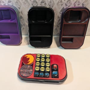 May include: Three rectangular plastic containers in purple, black, and brown. A red container with game pieces, including dice and tokens, is in the foreground. The game pieces have the numbers 100, 120, 60, and EX on them. The containers appear to be for a card game.