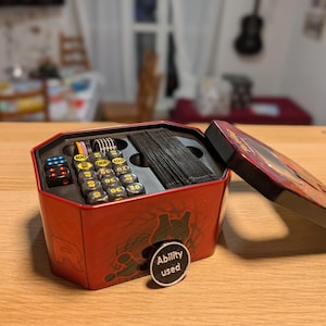 Könnte beinhalten: Eine rote, sechseckige Metalldose mit einem schwarzen Kartendeck, mehreren schwarzen Würfeln mit farbigen Punkten und schwarzen zylindrischen Spielsteinen mit gelben Zahlen. Ein runder schwarzer Spielstein mit dem Text Ability used ist befestigt.