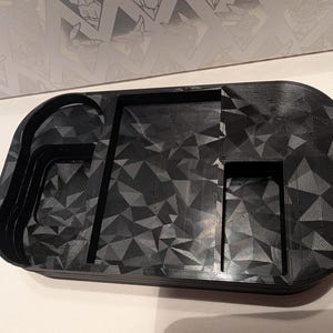 Könnte beinhalten: Eine schwarze Ablage mit geometrischem Muster und mehreren Fächern. Die Ablage hat eine abgerundete rechteckige Form mit strukturierter Oberfläche. Sie verfügt über einen Getränkehalter, einen rechteckigen Bereich und ein kleineres Fach.