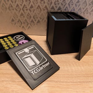 Könnte beinhalten: Schwarze, 3D-gedruckte Karten-Spiel-Aufbewahrungsbox mit abnehmbarem Deckel und einem separaten Tablett mit Spielmarken. Der Deckel trägt den Text "TCGärtner" und eine Grafik. Die Box steht auf einer Holzoberfläche.