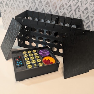 May include: Black 3D-printed game organiser with multiple compartments. Includes a tray with game tokens and dice, and a larger storage unit with dividers. The organiser has a geometric pattern on the sides and lid.