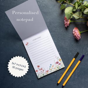 Könnte beinhalten: Ein personalisierter Notizblock mit 50 Seiten im A5-Format. Das weiße Papier hat horizontale Linien, eine Blumenillustration am unteren Rand, ein kleines Blumensymbol und den aufgedruckten Namen Lauren Jones. Zwei Stifte liegen neben dem Block.