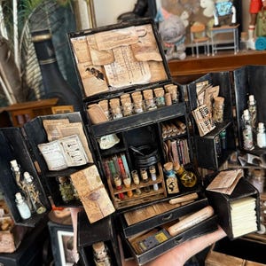 Könnte beinhalten: Eine Miniatur-Apothekerkiste in Schwarz, geöffnet, um Regale, Schubladen und Fächer mit winzigen Glasflaschen, Schriftrollen, Büchern und Phiolen zu zeigen. Die Kiste ist mit gealtertem Papier und Flaschen mit Korken verziert.