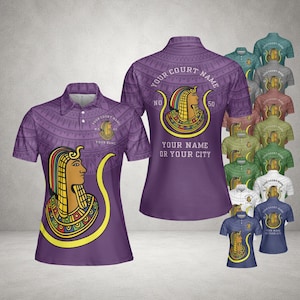 Peut inclure: Polo violet avec un motif égyptien. Le t-shirt présente un graphisme d'une figure égyptienne avec des accents dorés, jaunes et rouges. Le texte sur le t-shirt comprend "YOUR COURT NAME", "YOUR NAME OR YOUR CITY" et "NO 50". Plusieurs autres variations de couleurs sont également affichées.