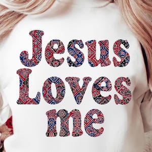 Pode incluir: Camisola branca com a frase "JESUS LOVES me" num padrão decorativo vermelho, azul e preto. O texto está centrado na frente da camisola.