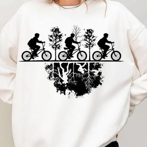 May include: White sweatshirt featuring a black graphic of three cyclists riding bicycles past trees. Below, a mirrored image of the scene depicts an upside-down world with roots and abstract shapes. The design is a silhouette.