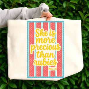 Puede incluir: Bolso tote color crema con un diseño rectangular. El diseño presenta el texto "She is more precious than rubies" en escritura amarilla. El diseño tiene un fondo a rayas rojas y blancas con lunares azules y un borde festoneado azul. El texto "PROV 3:15" está en la parte inferior.