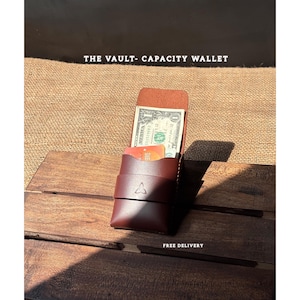 Peut inclure: Un portefeuille en cuir marron foncé avec le texte "THE VAULT- CAPACITY WALLET". Le portefeuille est ouvert, révélant un billet d'un dollar et une carte de crédit. Le portefeuille a une sangle avec un logo triangulaire.
