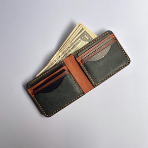 May include: A bifold wallet featuring dark charcoal grey leather pockets and a tan leather interior. The wallet is constructed with thick, visible orange stitching along the edges and contains several United States one-dollar bills.