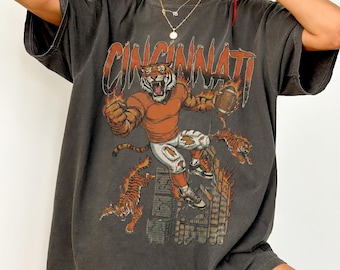 Può includere: T-shirt oversize grigio scuro con grafica a tigre dei Cincinnati Bengals. La grafica include la parola "Cincinnati" in arancione, una tigre in uniforme da football e ulteriori illustrazioni di tigri. La maglietta ha maniche corte.