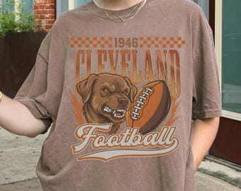 May include: Brown t-shirt with a graphic of a dog and a football, and the text "Cleveland Football 1946". The shirt is short-sleeved and has a crew neck.