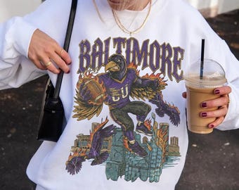 May include: White sweatshirt featuring a raven graphic holding a football, with the word "BALTIMORE" above. The raven is in a purple jersey. A brown baseball cap with "La Poésie PARIS" is worn.