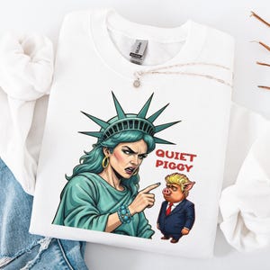 May include: White sweatshirt featuring a graphic of the Statue of Liberty pointing at a cartoon pig wearing a suit. The text "QUIET PIGGY" is in red. The Statue of Liberty is depicted with green skin and a stern expression.